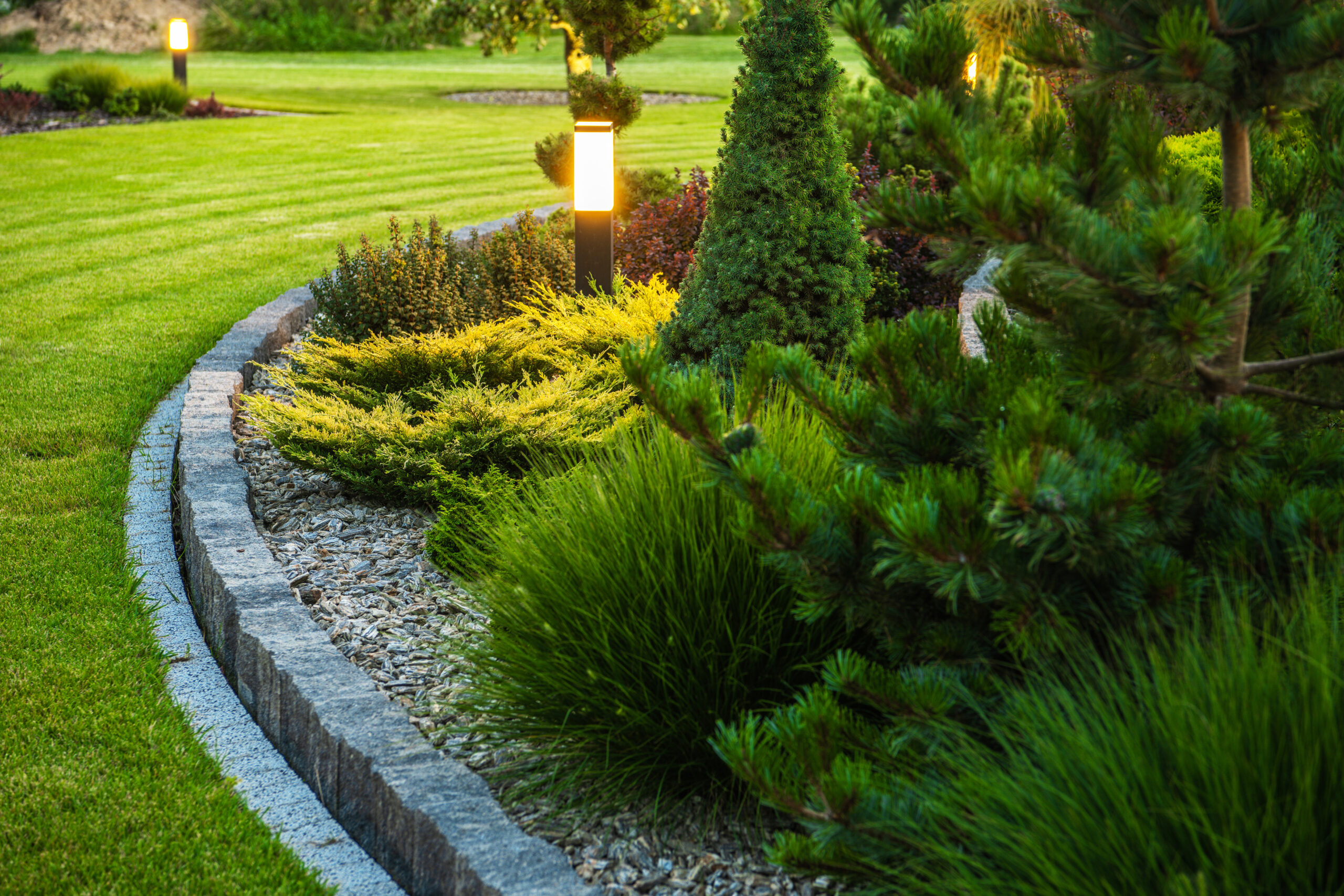 Landscaped Garden at the Backyard with Evenly Mowed Lawn and Island Bed with a Variety of Shrubs, Bushes, Trees and Evergreen Plants. Decorated with Bollard Outdoor Lighting Lamp.